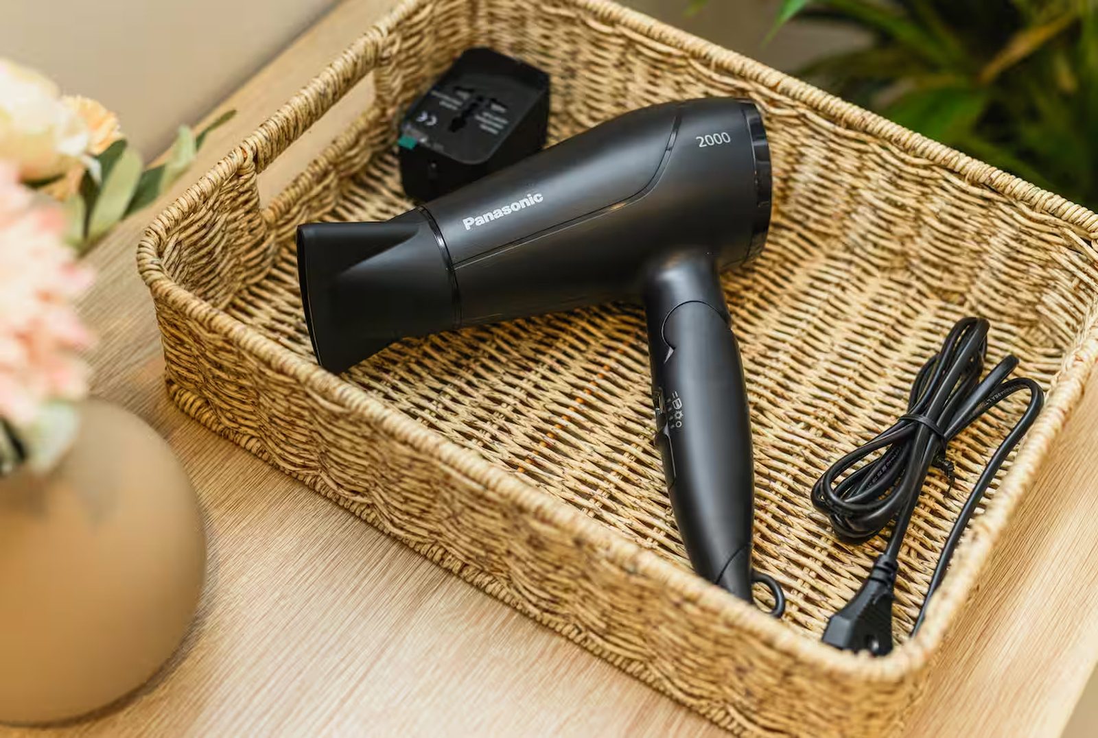 A woven basket on a wooden surface holding a neatly coiled hairdryer, beside a vase of flowers